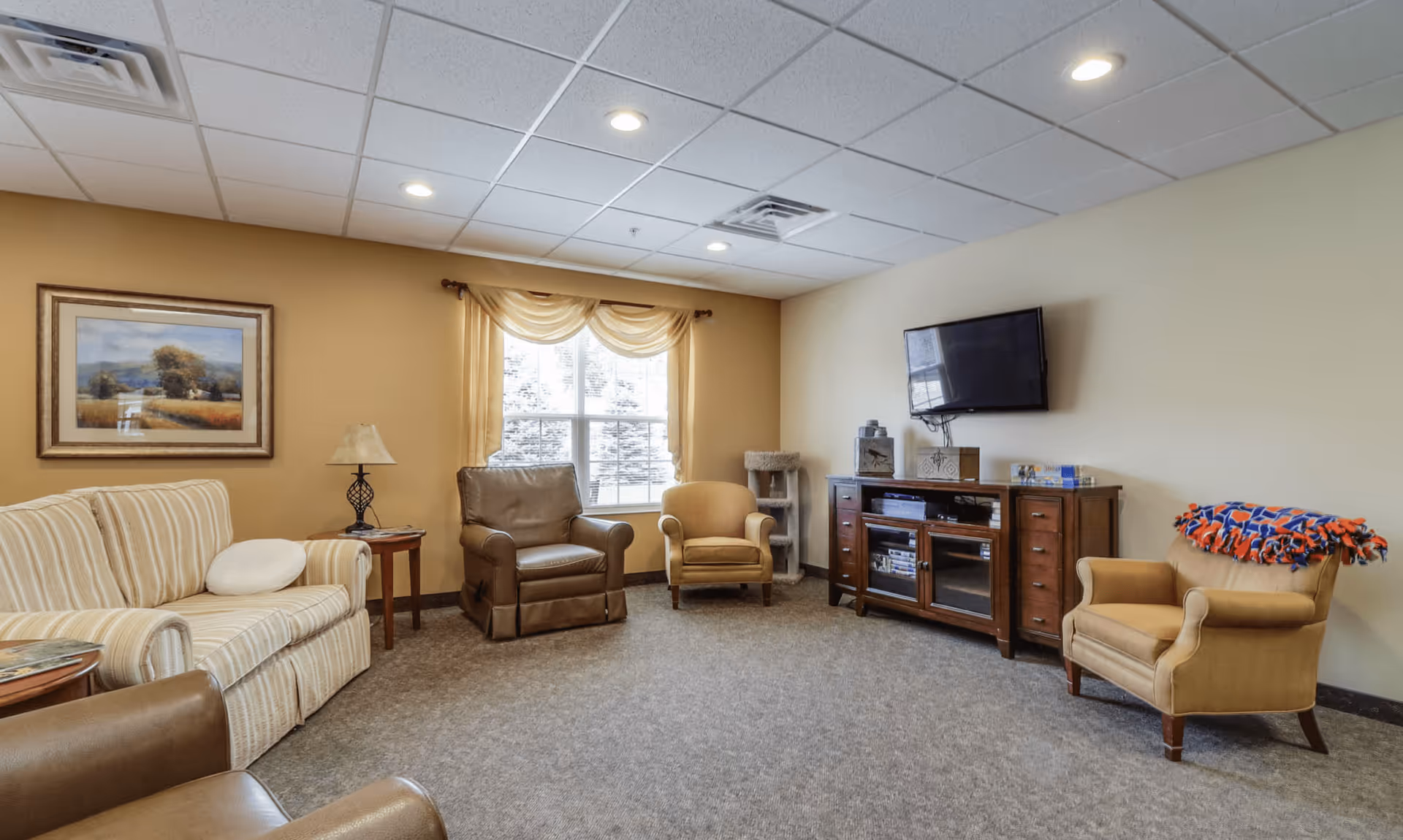 A cozy living room with beige walls and carpeted floor, featuring a striped sofa, three armchairs, a wooden side table with a lamp, a window with light curtains, a wall-mounted flat screen TV above a wooden media cabinet, and a colorful blanket draped over one armchair.