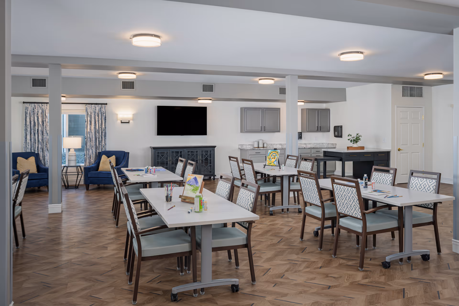 A bright communal dining and activity room with multiple tables and chairs, a wall-mounted TV, kitchenette, and lounge seating.