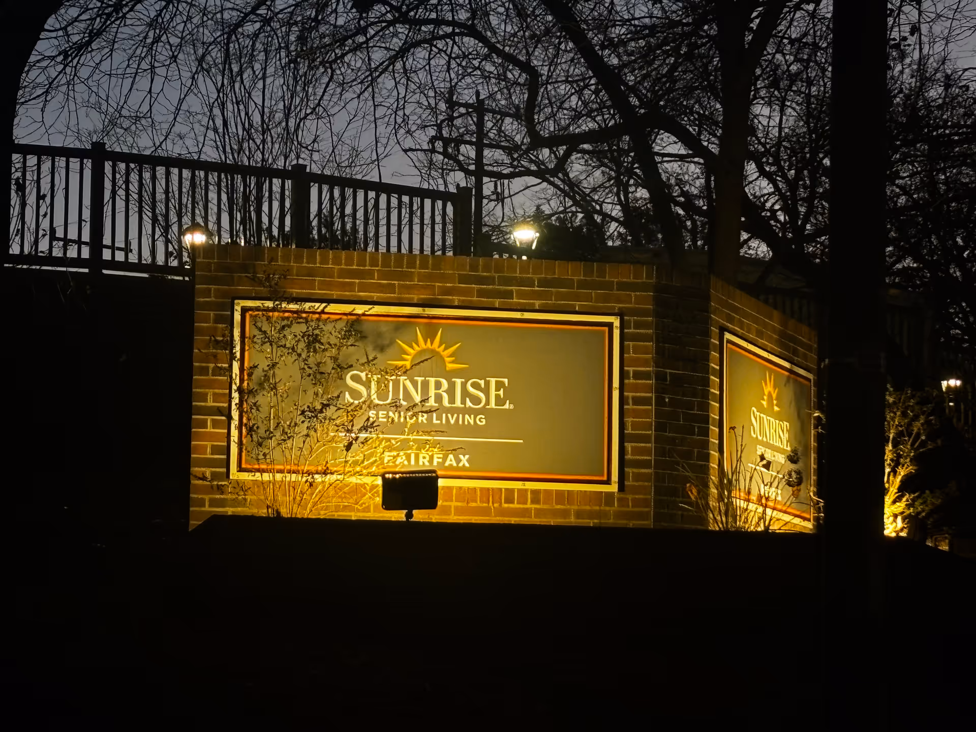 Illuminated brick sign for Sunrise Senior Living Fairfax at night, surrounded by leafless trees and a metal fence in the background.