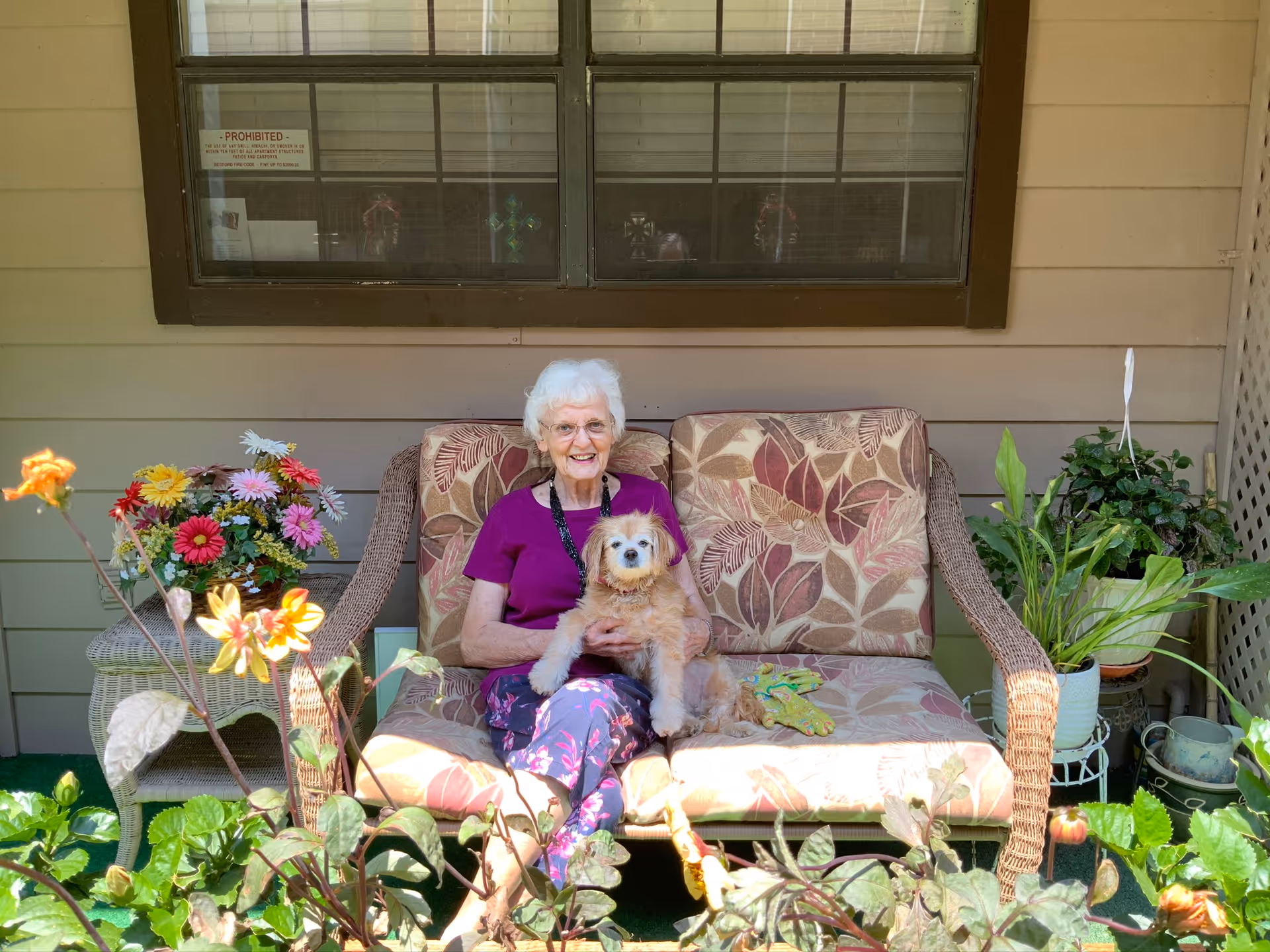 An elderly woman with white hair and glasses sitting on a cushioned outdoor wicker loveseat, holding a small fluffy dog. The setting is a porch or patio area with potted plants and flowers around, and a window with blinds behind her.
