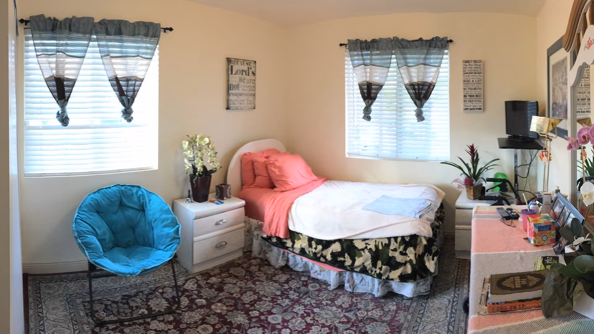 Bright bedroom with a single bed with pink bedding, nightstands, a turquoise accent chair, dresser and plants beneath two windows.