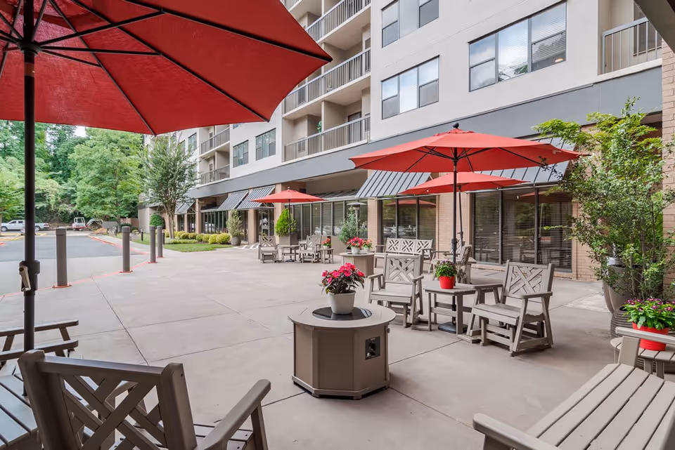 Outdoor patio area at Hammond Glen Retirement Community with multiple seating arrangements including wooden chairs and tables under large red umbrellas. Potted plants with flowers decorate the space, and the building with large windows and balconies is visible in the background.