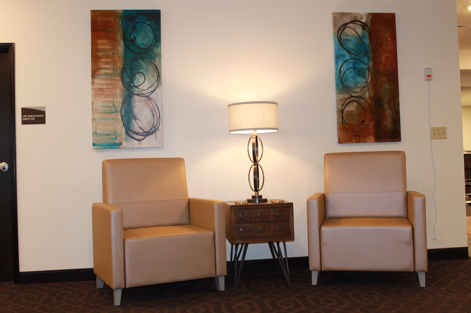 Two tan armchairs separated by a small wooden side table with a modern lamp on it, positioned against a beige wall with two abstract paintings hanging above each chair. A door with a sign reading 'Life Enrichment Director' is visible on the left side.