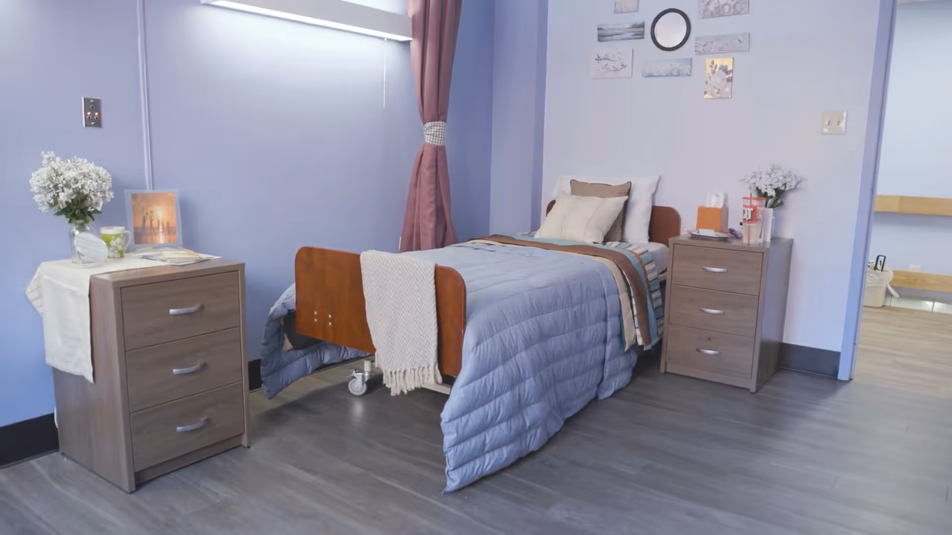 A neatly made single bed with a blue quilt and multiple pillows in a room with light purple walls. There are two wooden bedside cabinets on either side of the bed, each decorated with a vase of white flowers and other small items. A maroon curtain is tied back on the window behind the bed, and several small framed pictures and a round mirror are hung on the wall above the bed. The floor is covered with gray wood-like flooring, and an open doorway leads to another room with light-colored flooring.