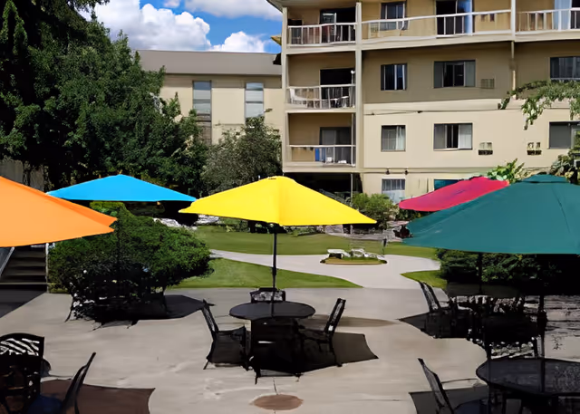 Outdoor patio area with multiple round tables and colorful umbrellas in orange, blue, yellow, pink, and green. The patio is surrounded by greenery and bushes, with a multi-story beige building in the background under a partly cloudy sky.