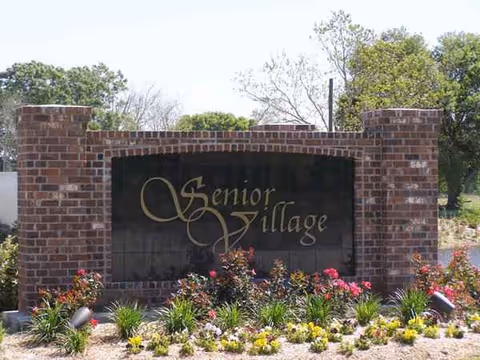 Brick entrance sign reads Senior Village, surrounded by colorful flowers and greenery.
