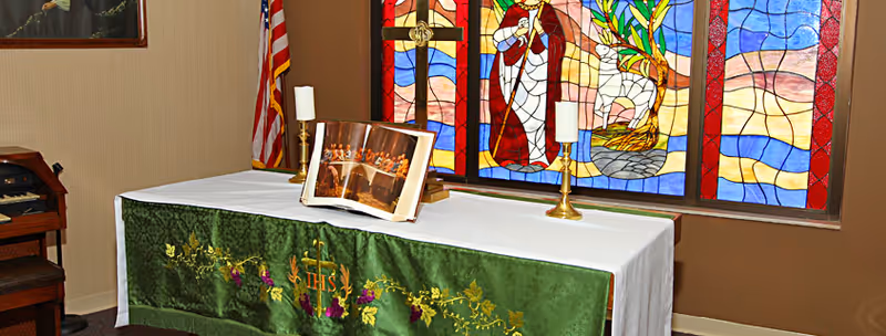 An altar inside a room with a white cloth and a green embroidered altar frontal. On the altar are two candlesticks with white candles and an open book displaying a religious image. Behind the altar is a colorful stained glass window depicting religious figures and symbols. An American flag is positioned to the left side of the altar.