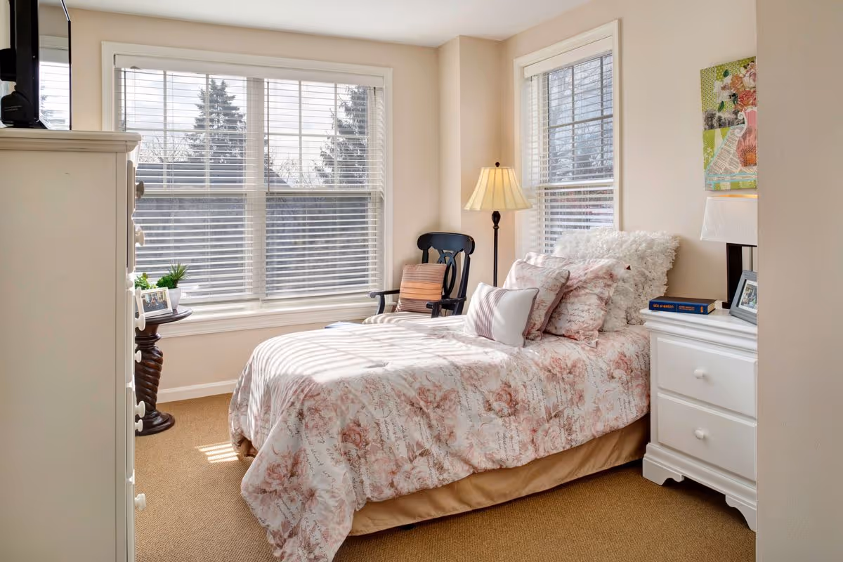A cozy bedroom with a single bed covered in a floral patterned comforter and multiple pillows. The room has beige walls and carpet, two large windows with white blinds letting in natural light, a black wooden chair with a cushion, a floor lamp, a white dresser with a table lamp, books, and framed photos on top, and a small round table with a plant and photo frame near the window.