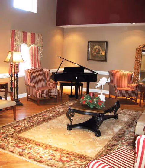 Cozy senior living lounge with striped chairs, a grand piano, lamp, and decorative rug.