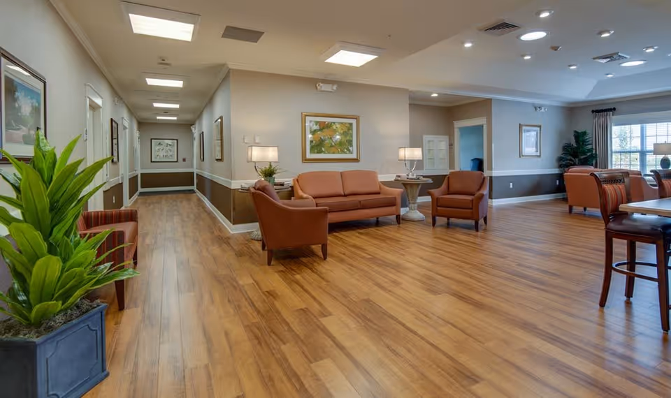 A spacious senior living facility common area with wooden flooring, several orange upholstered chairs and sofas, side tables with lamps, framed artwork on the walls, large windows with curtains, and green plants in decorative pots.