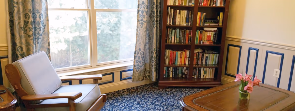 A cozy sitting area with a cushioned wooden armchair next to a large window with patterned curtains. There is a wooden bookshelf filled with books and a wooden coffee table with a small vase of pink flowers on it. The room has blue and white patterned carpet and decorative wall paneling.