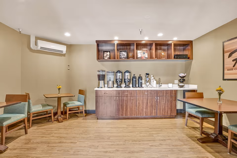 A small dining area with wooden tables and chairs featuring green cushions. Against the back wall is a wooden cabinet with a white countertop holding several beverage dispensers, cups, and a sink. Above the cabinet are wooden shelves displaying framed pictures and decorative items. The room has beige walls, wooden flooring, and recessed ceiling lights.