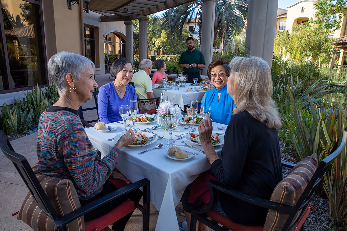 A group of elderly people sitting at two outdoor dining tables covered with white tablecloths, enjoying a meal together under a pergola with greenery and palm trees in the background. A man in a green shirt stands near the tables, possibly serving or talking to the group.