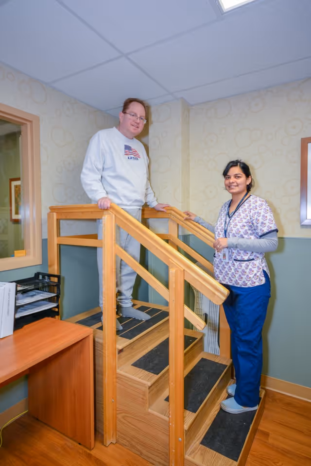 A man in casual clothing stands on a small wooden staircase with handrails inside a room, while a healthcare worker in scrubs stands beside the stairs smiling. The room has light-colored walls with a circular pattern wallpaper and a wooden floor.