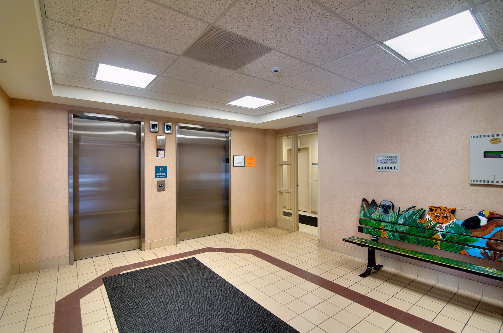 Interior view of a senior living facility elevator lobby with two stainless steel elevators, beige walls, tiled floor with a dark mat, and a colorful bench painted with jungle animals including a tiger, toucan, and other birds. There is a door leading to another room on the right side.
