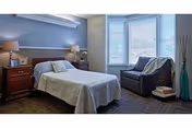 Sunlit bedroom with a double bed flanked by nightstands and lamps, and a cushioned armchair by a bay window.