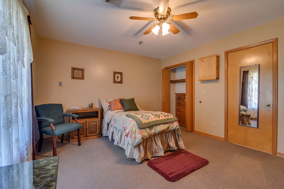 A cozy bedroom in a senior living facility with a single bed covered in a floral quilt and pillows. There is a wooden nightstand with a telephone and a green upholstered chair next to it. The room has beige walls, a ceiling fan with lights, a closet with wooden doors, and a door with a full-length mirror. Sheer curtains cover a window on the left side, allowing natural light to enter.