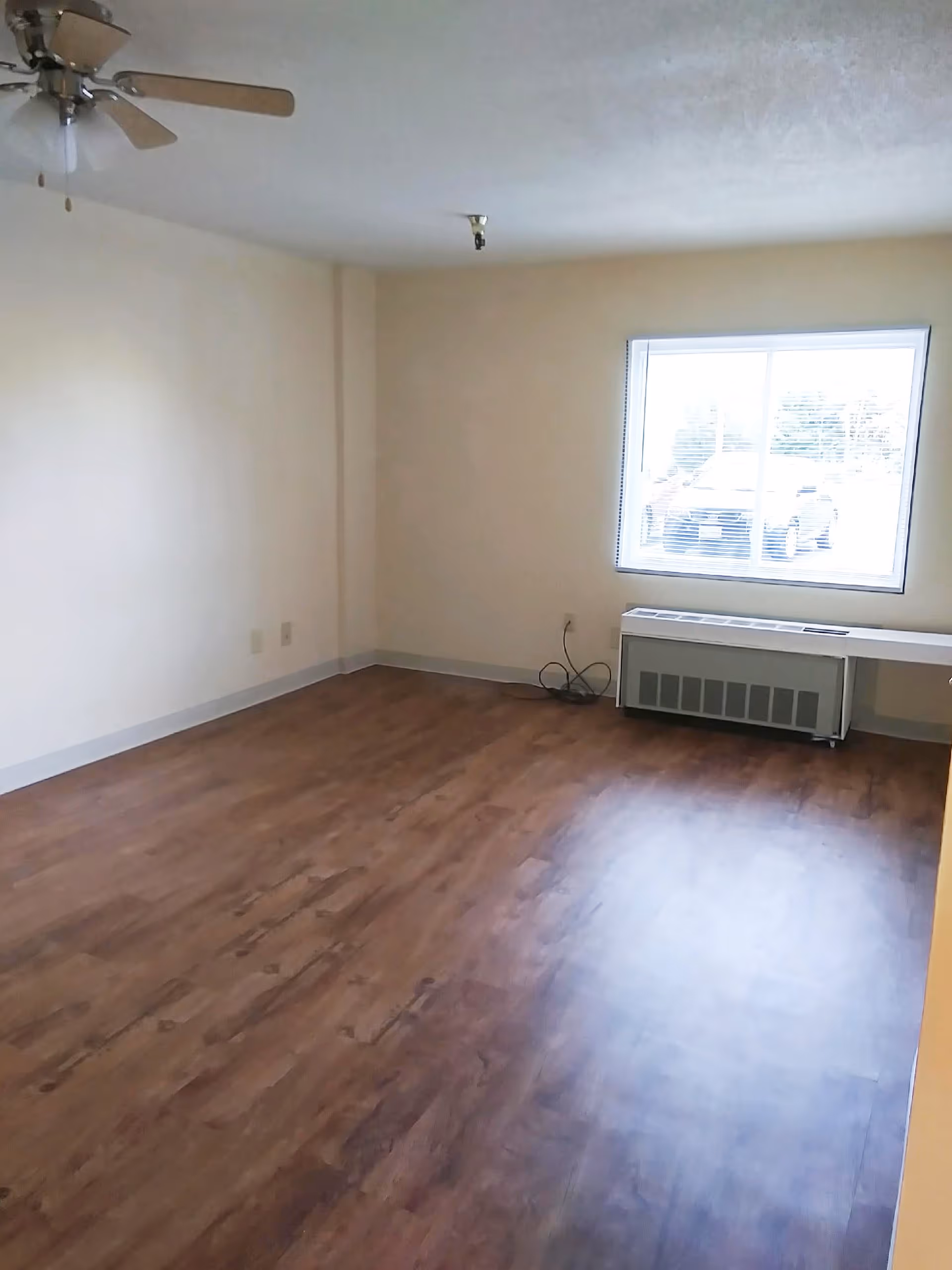 Empty room with wooden flooring, beige walls, a ceiling fan with wooden blades, a large window with blinds, and a heating or air conditioning unit below the window.