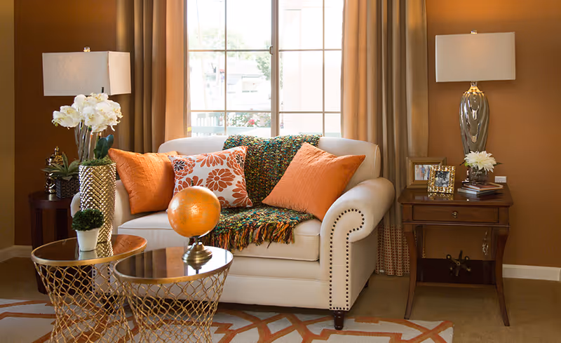 Cozy living room with a beige loveseat topped with orange and patterned pillows and a colorful throw, flanked by side tables, lamps, and decorative accents in front of a window.