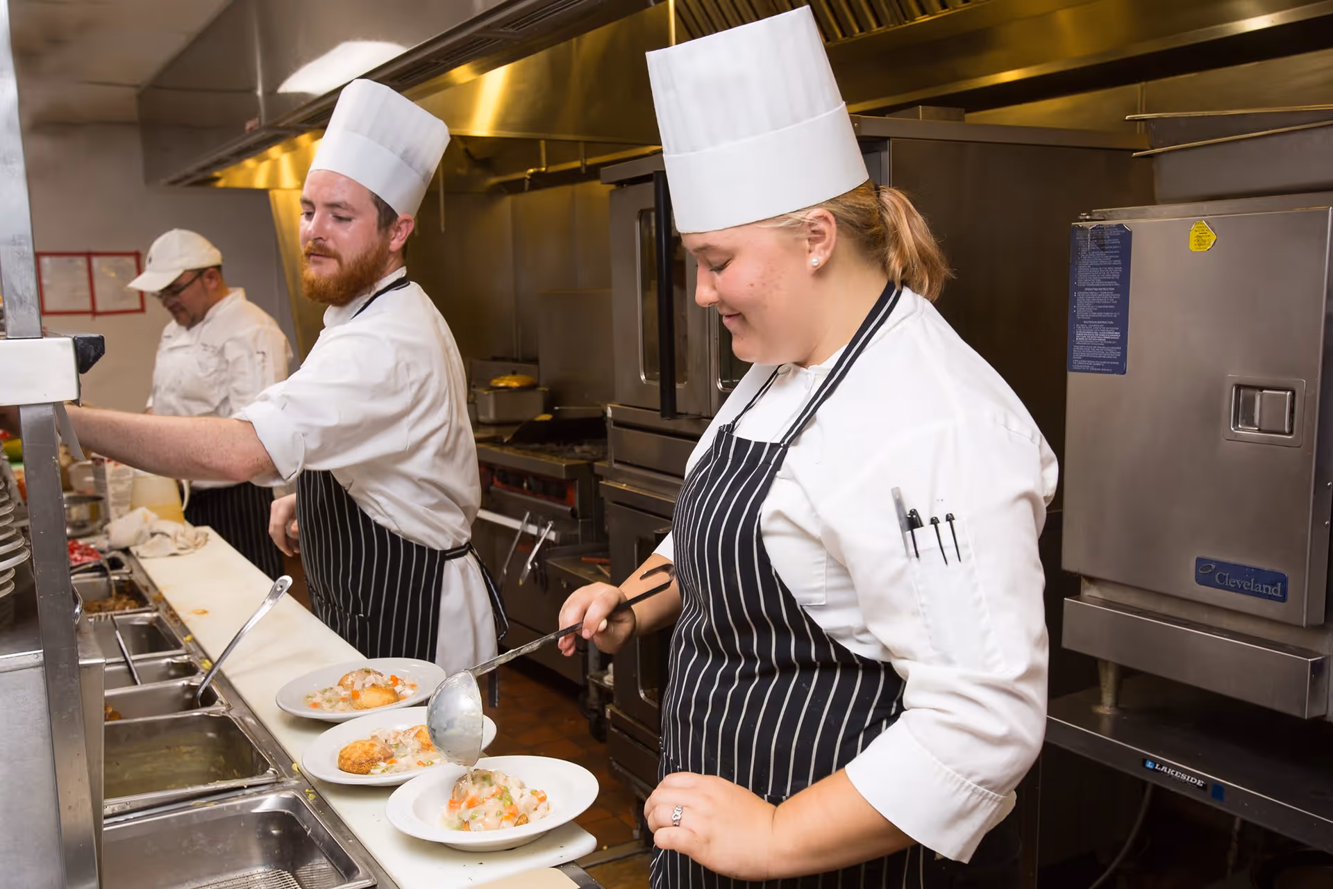 Two chefs wearing white uniforms and tall white hats are preparing and plating food in a commercial kitchen. One chef is ladling a stew or soup onto a plate, while the other is reaching for something on the counter. A third person in a white cap is working in the background.