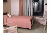 A simple bedroom in a care facility featuring a single bed with a pink bedspread and a teddy bear placed on it. The room includes two light pink armchairs, a window with vertical blinds, and a small wall-mounted light fixture.