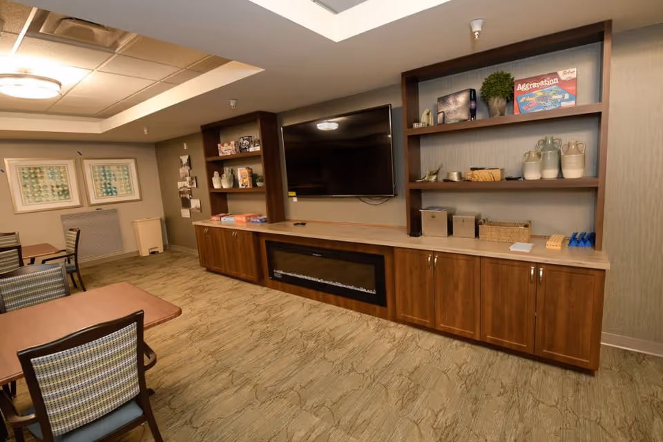 A common area in a senior living facility featuring a large flat-screen TV mounted on the wall above a wooden cabinet with an electric fireplace. The cabinet has shelves with decorative items, board games, and storage boxes. There are tables and chairs with patterned upholstery arranged in the room, and framed artwork is displayed on the wall.