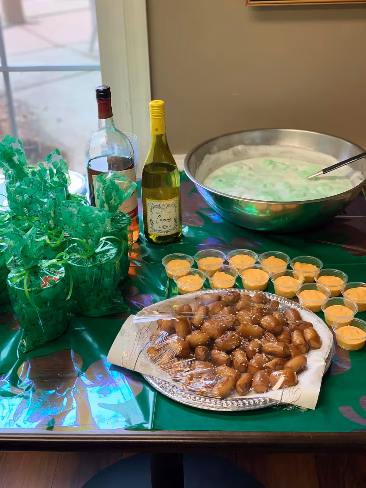 Table with a tray of bite-sized pastries, small cups of dipping sauce, two wine bottles, green-wrapped favors, and a large bowl of green punch.