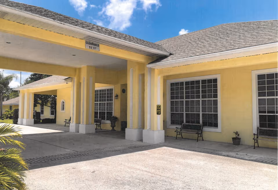 Front entrance of a yellow building with a covered porte-cochere, large windows and benches.