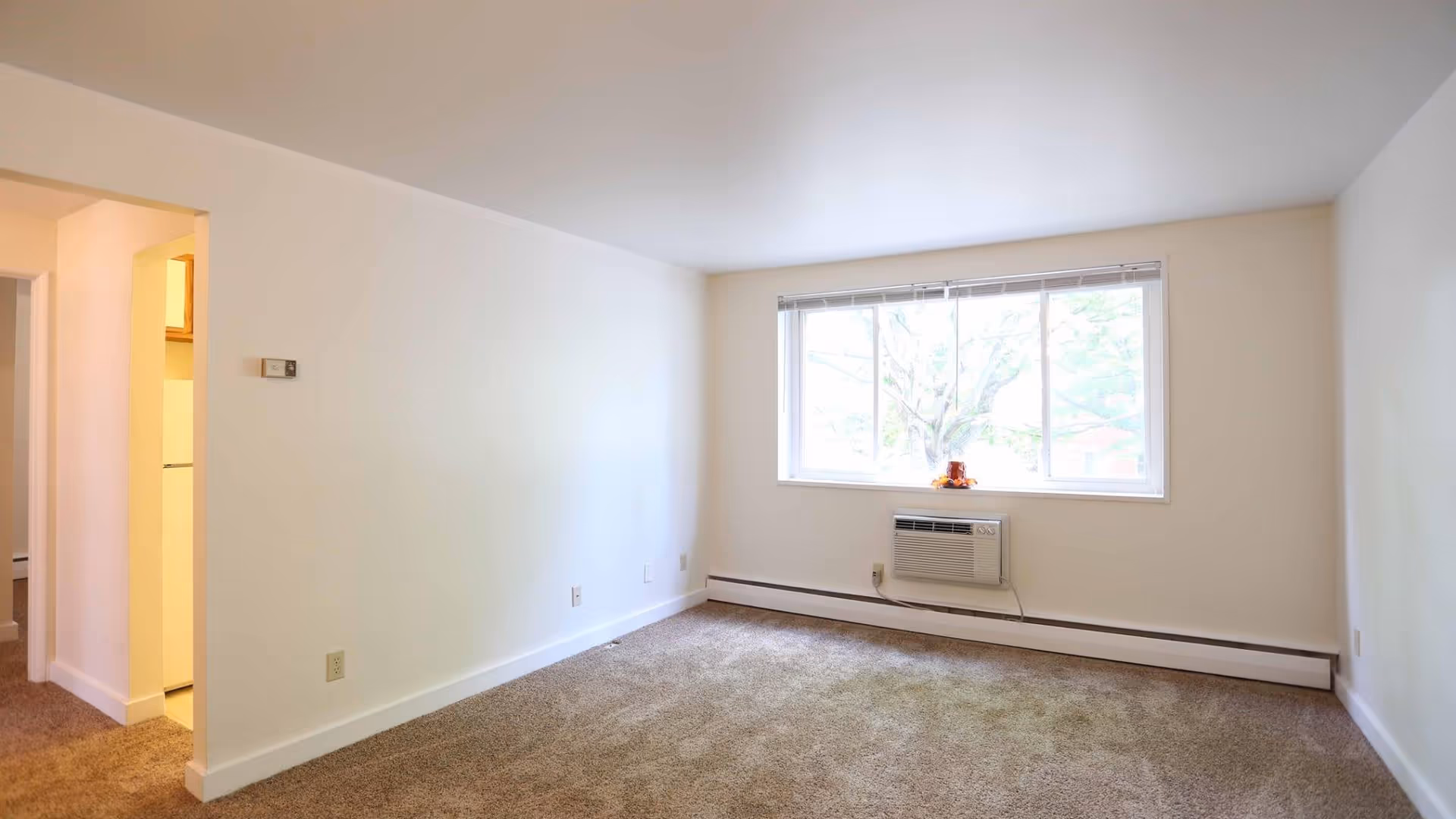 Empty room with beige carpet, white walls, a large window with blinds, and a wall-mounted air conditioning unit below the window. There is an open doorway leading to a kitchen area with a refrigerator visible.