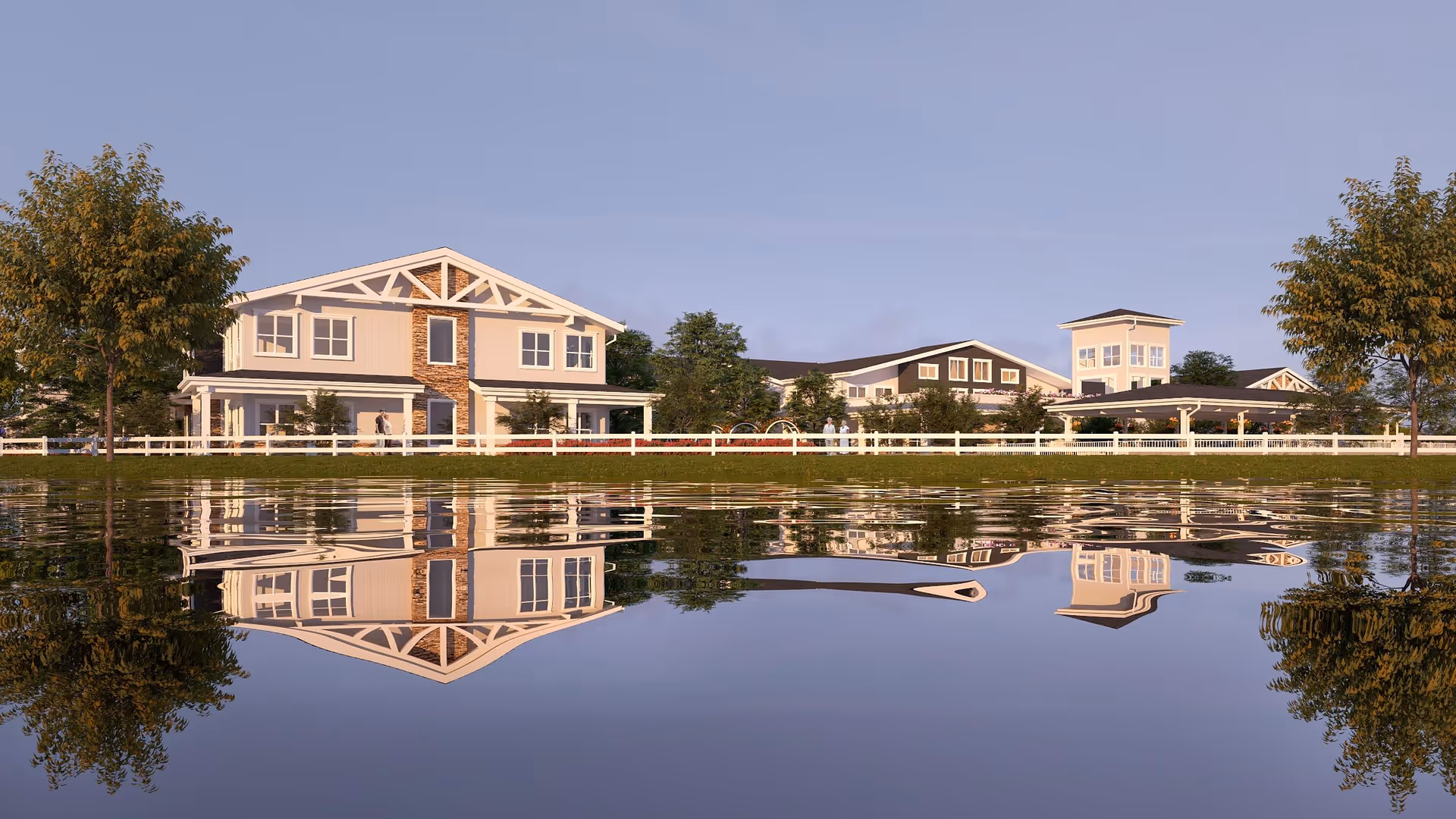 A serene view of The Watermark at Morrison Ranch facility showing a large building with white and beige exterior, surrounded by trees and a white fence, reflected in a calm body of water in the foreground under a clear sky.