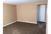 Empty room with beige walls, brown carpeted floor, white baseboards, and a white door slightly open leading to another room.