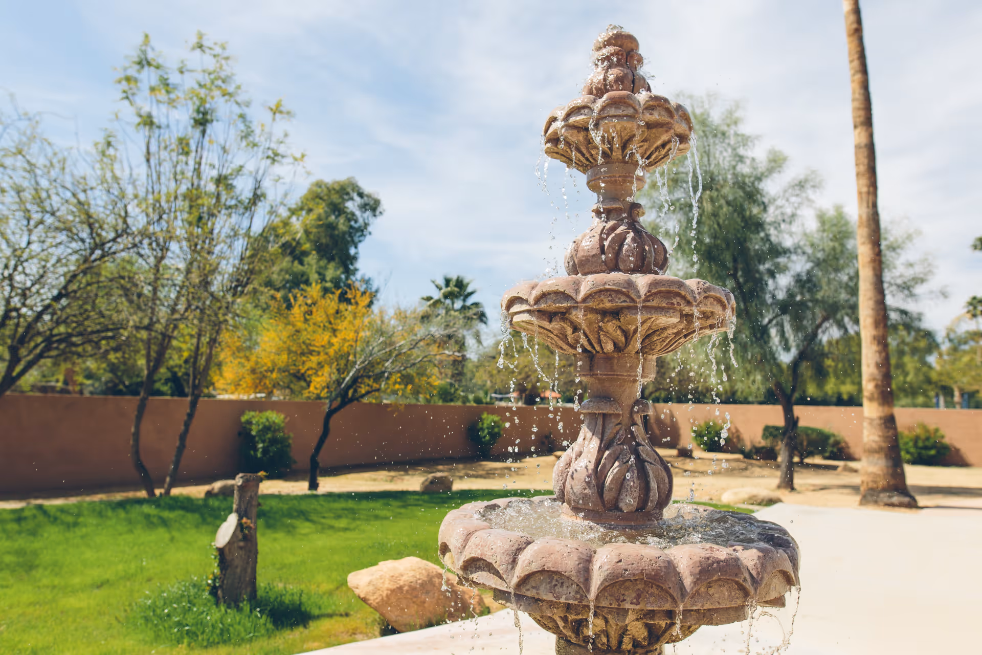 A three-tier stone fountain spraying water in a landscaped courtyard with trees and a low wall in the background.