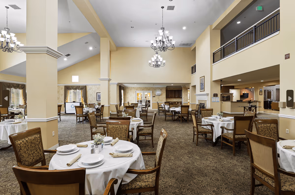 Spacious dining room in an assisted living facility with multiple round tables covered with white tablecloths, set with plates, napkins, and utensils. The room features high ceilings, chandeliers, beige walls, patterned carpet, and large windows with curtains. There is a fireplace and a balcony overlooking the dining area.