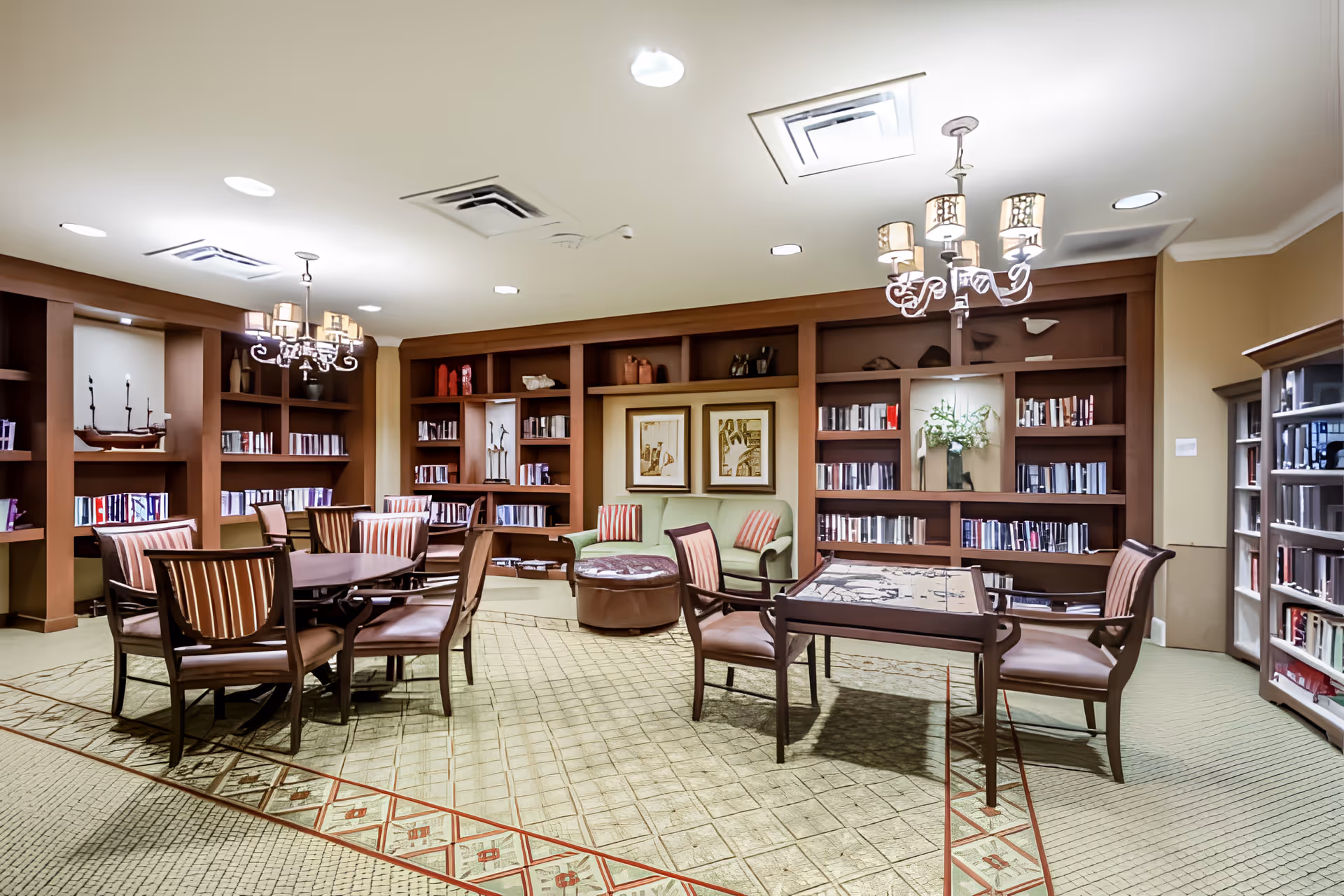 A well-lit senior living community library and lounge area featuring wooden bookshelves filled with books, two chandeliers, multiple tables with chairs, a green armchair with striped cushions, and framed artwork on the wall.
