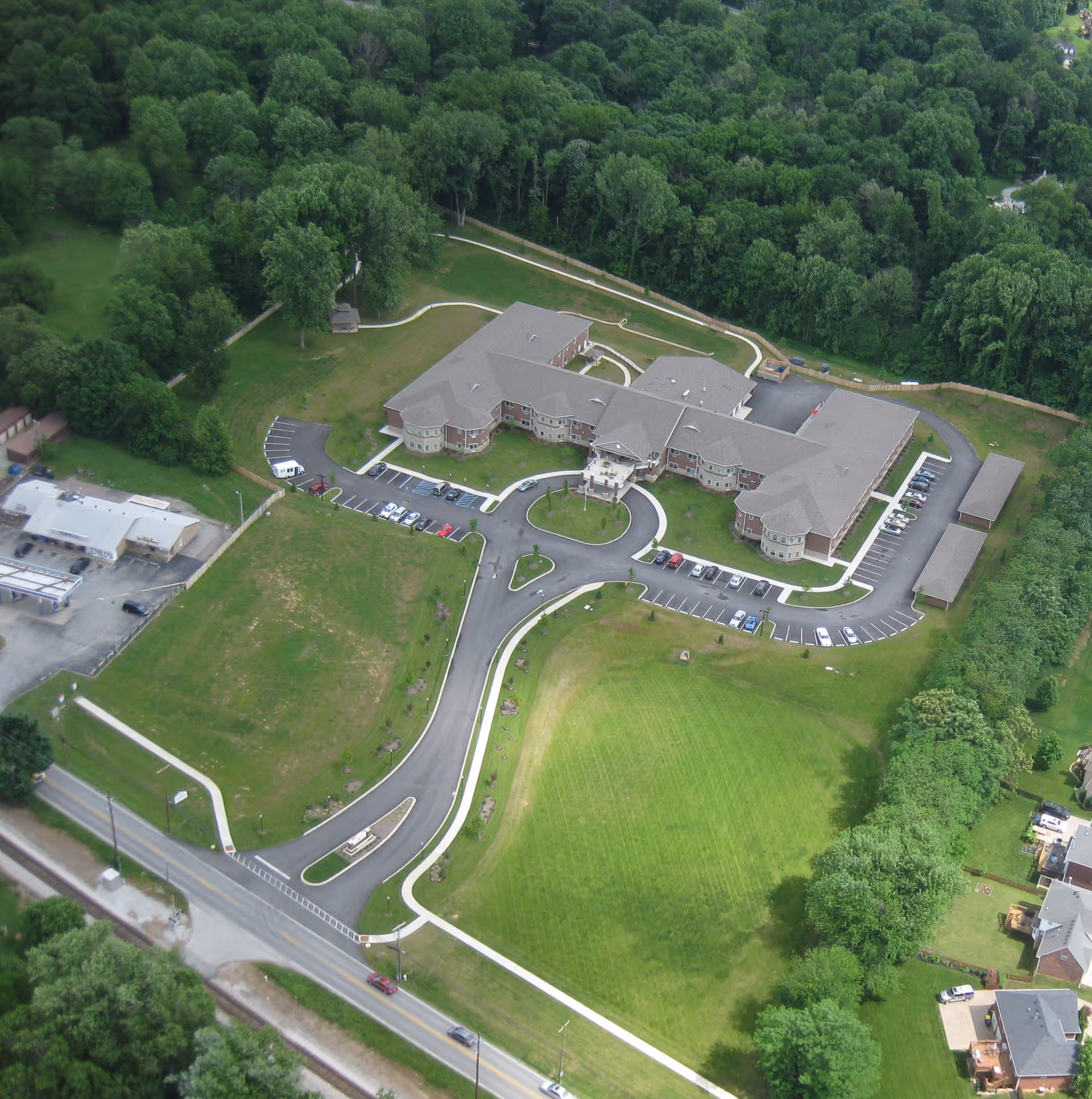 Aerial view of Magnolia Springs East Louisville facility surrounded by green trees and lawns. The building is large with multiple wings and a circular driveway at the entrance. There are several parking spaces with cars parked, and a road leading to the facility. The surrounding area includes wooded regions and some residential houses.