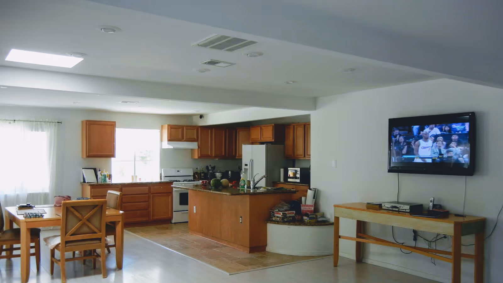 Open-plan kitchen and dining area with wooden cabinets, a central island, dining table with chairs, and a wall-mounted TV showing a basketball game.