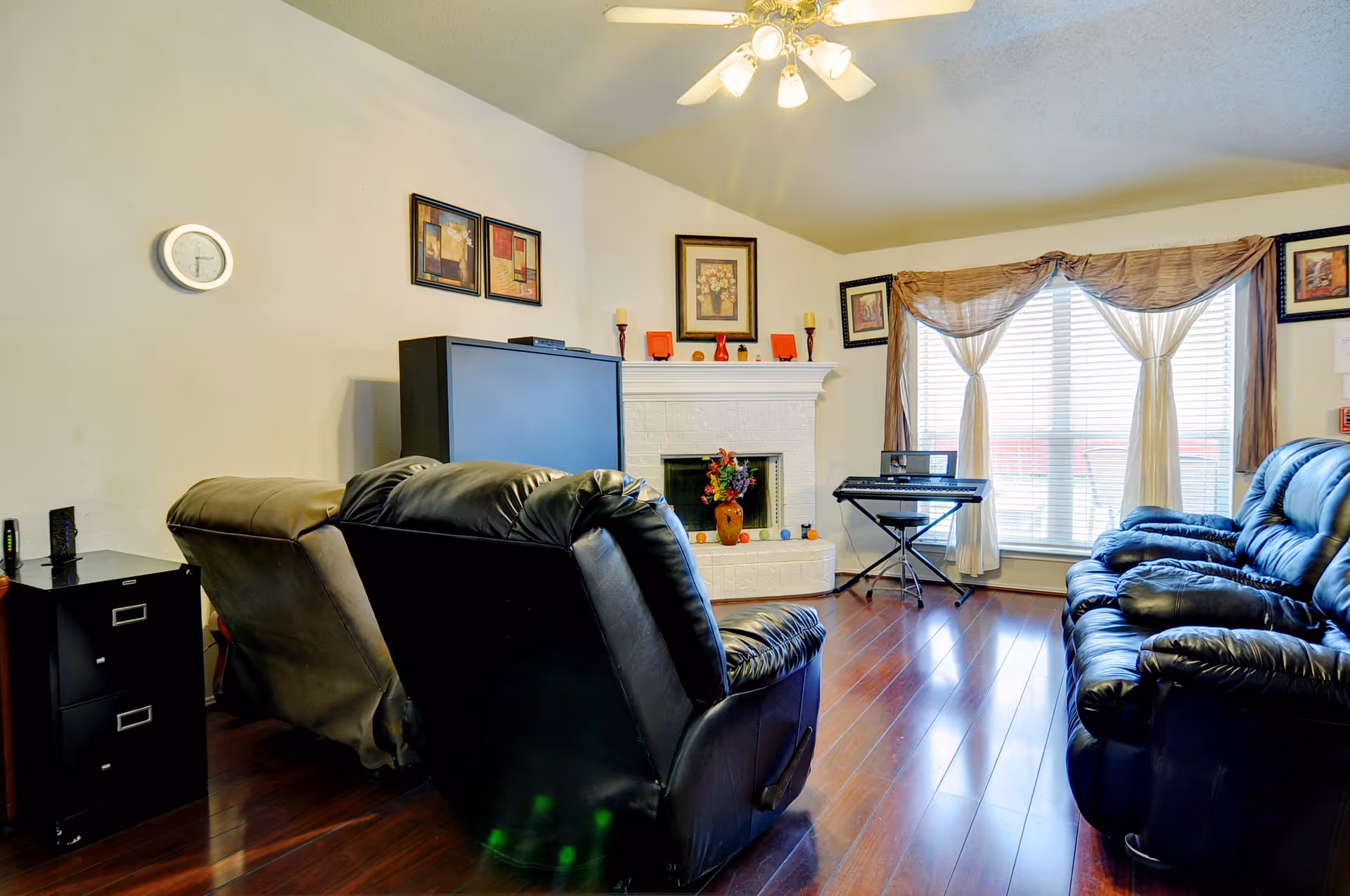 A cozy living room with dark leather recliner chairs and a sofa arranged around a white brick fireplace. Above the fireplace are framed pictures and decorative candles. A large window with sheer curtains allows natural light to fill the room. A keyboard on a stand is placed near the window, and a flat-screen TV is positioned on a stand against the wall. The room has wooden flooring and a ceiling fan with lights.