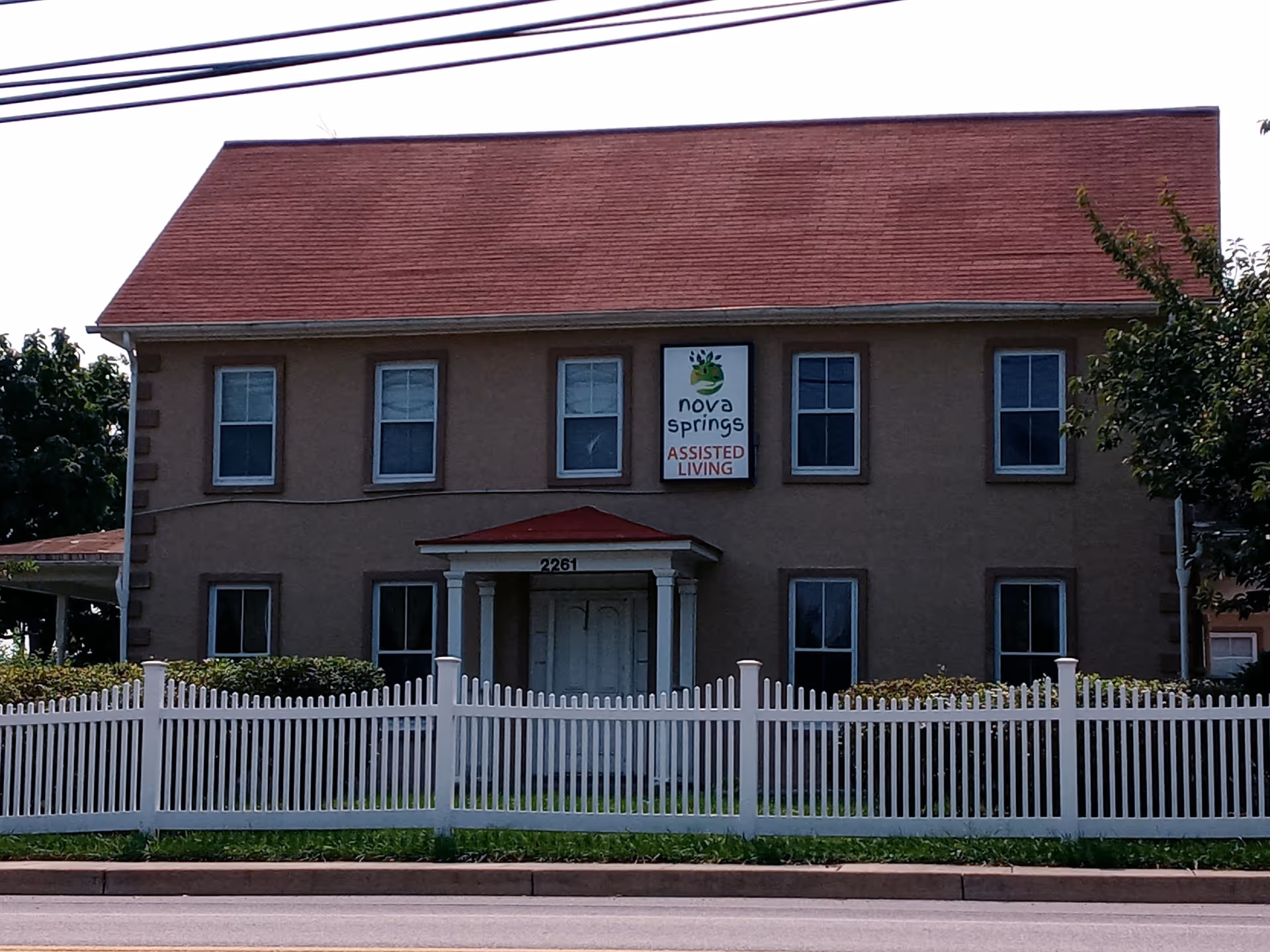 Exterior view of a two-story building with a red roof and beige walls, featuring a white picket fence in front. The building has six windows visible and a central entrance with a small porch. A sign above the entrance reads 'nova springs ASSISTED LIVING'.