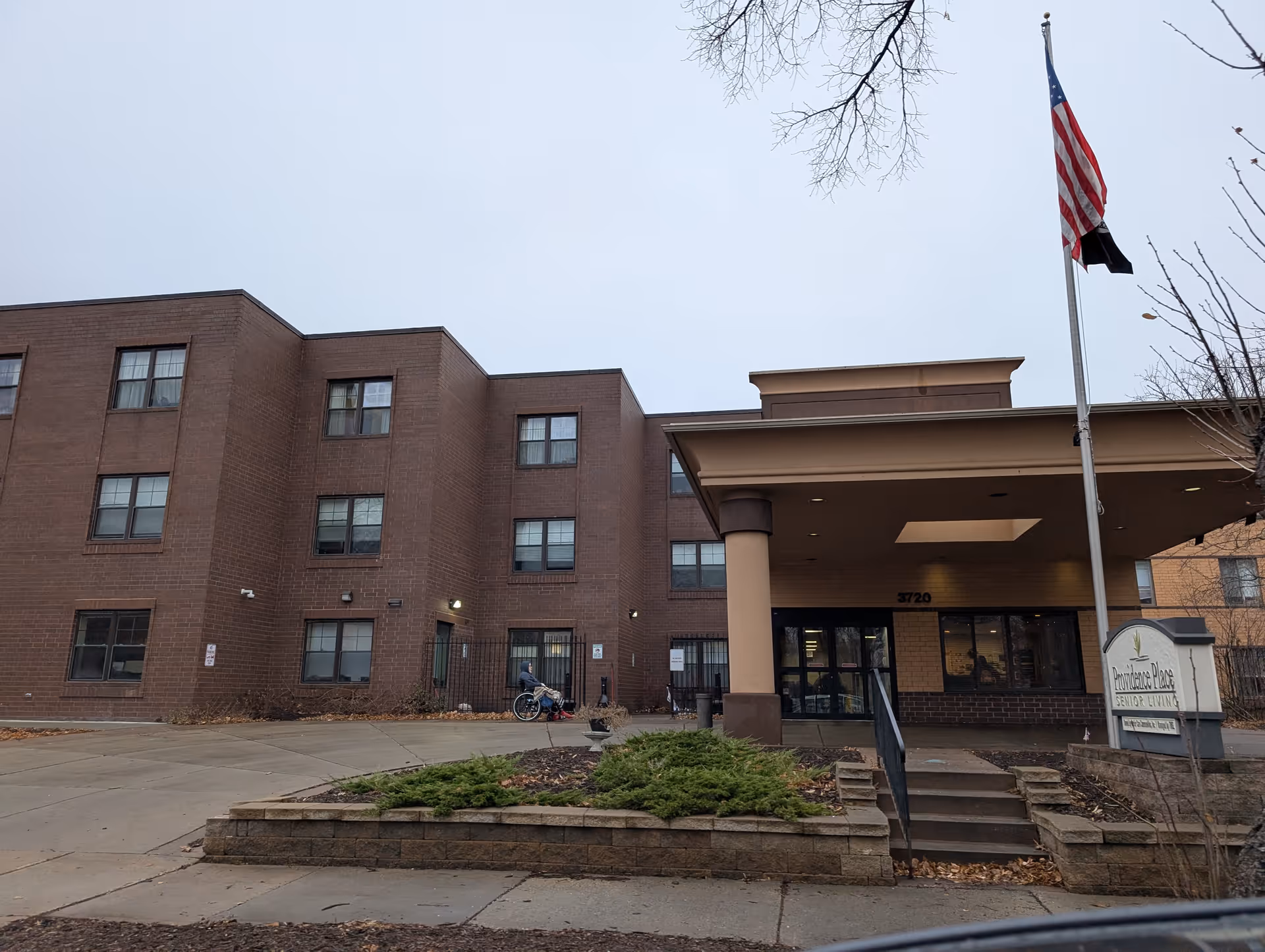 Exterior view of a three-story brick building with an entrance canopy supported by columns. A person in a wheelchair is near the entrance. There is a flagpole with an American flag and a sign that reads 'Providence Place Senior Living'. The sky is overcast and there are some bare tree branches visible.