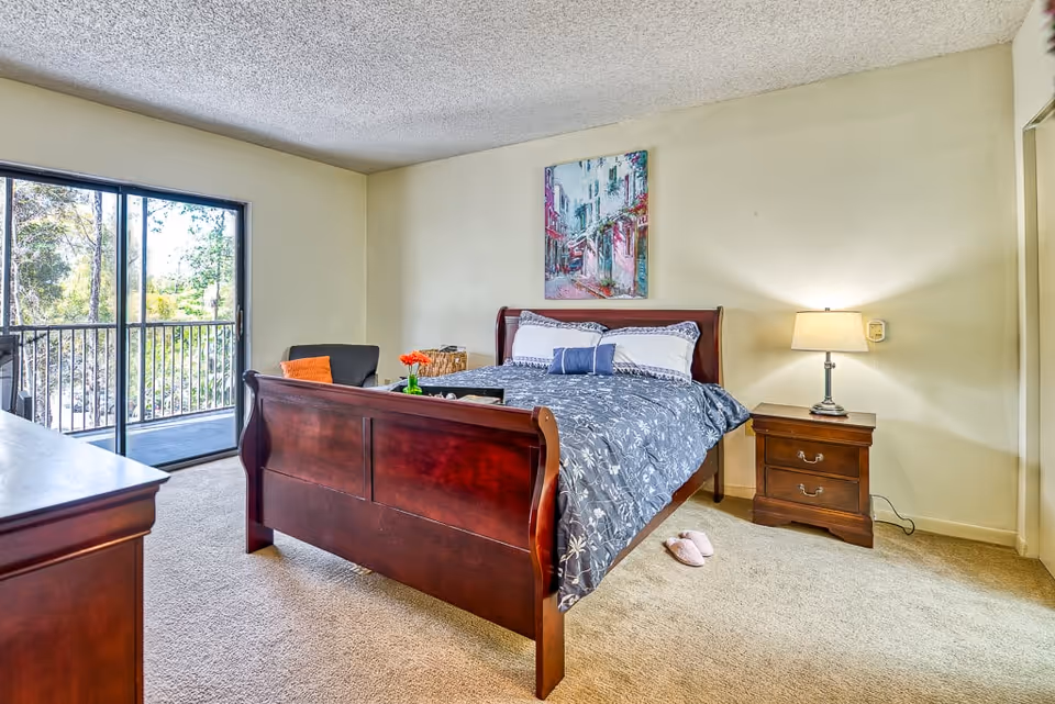 A bright bedroom with a wooden bed frame featuring a blue and white floral comforter and pillows. There is a wooden nightstand with a lamp on the right side of the bed and a wooden dresser on the left. A sliding glass door leads to a balcony with a view of trees outside. A colorful painting hangs on the wall above the bed, and a pair of pink slippers are on the carpeted floor near the bed.
