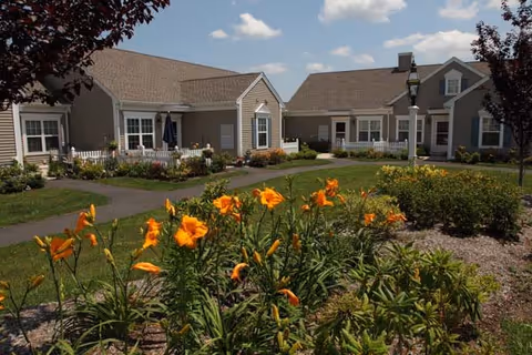 View of a residential community with single-story beige buildings, a well-maintained garden featuring orange flowers in the foreground, paved walkways, and a clear blue sky with a few clouds.