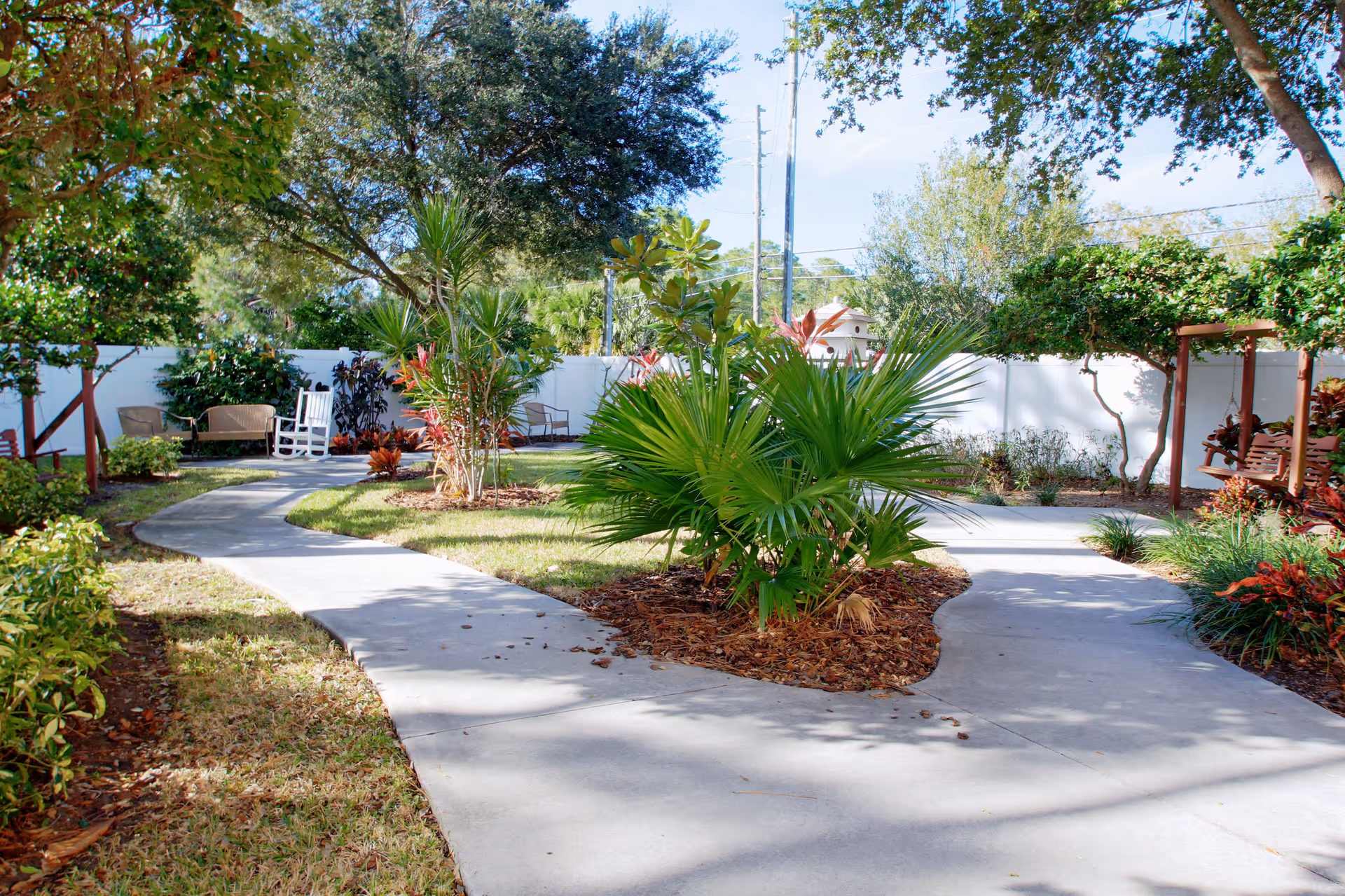 A landscaped outdoor garden area with a winding concrete pathway surrounded by green plants, trees, and shrubs. There are benches and a white rocking chair along the path, with a white fence enclosing the space.