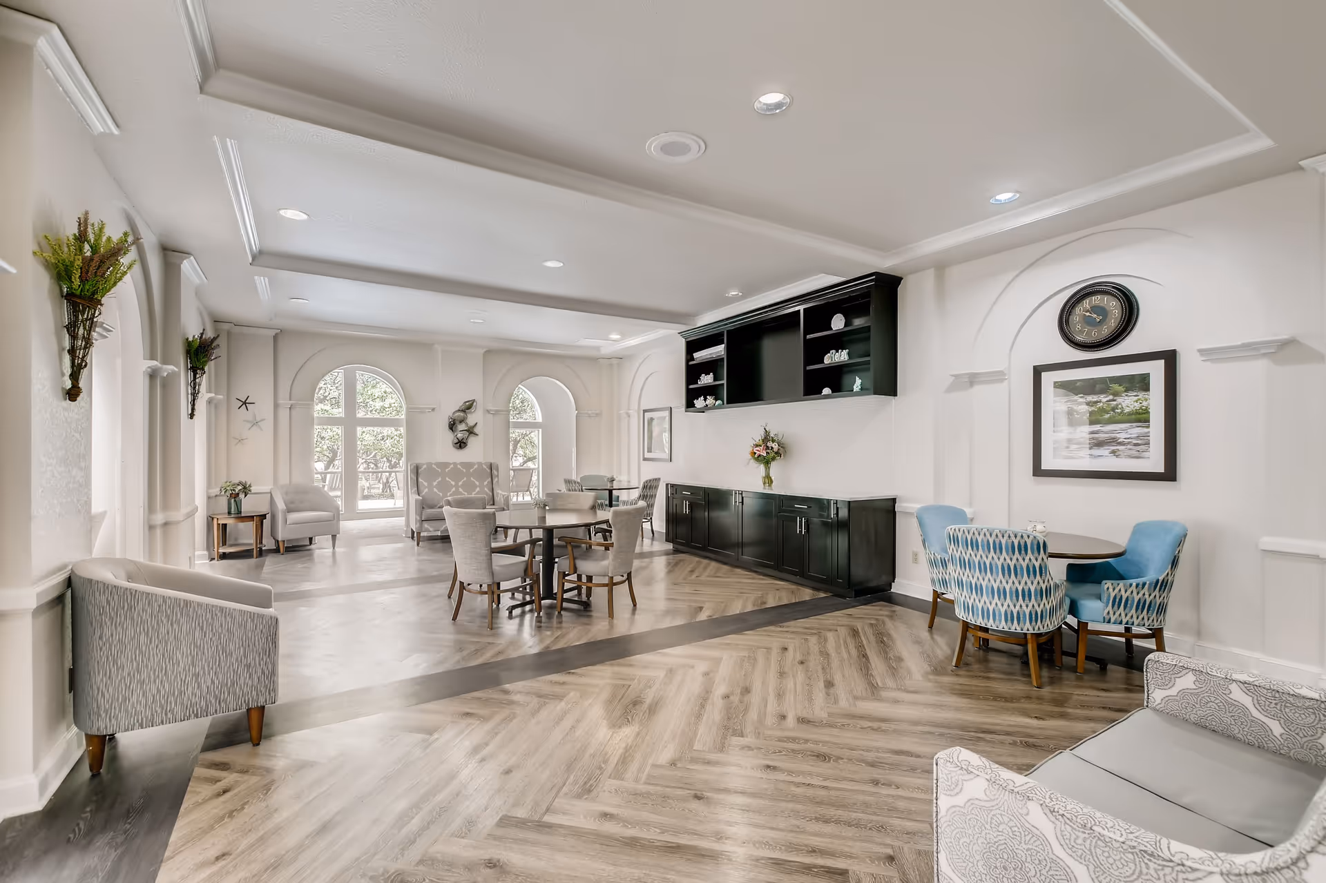 A bright and spacious senior living common area with multiple seating arrangements including round tables with chairs, armchairs, and a loveseat. The room features large arched windows letting in natural light, decorative wall sconces with greenery, a wall clock, framed artwork, and a built-in black cabinet with shelves displaying decorative items. The flooring is a light wood pattern with a darker border.