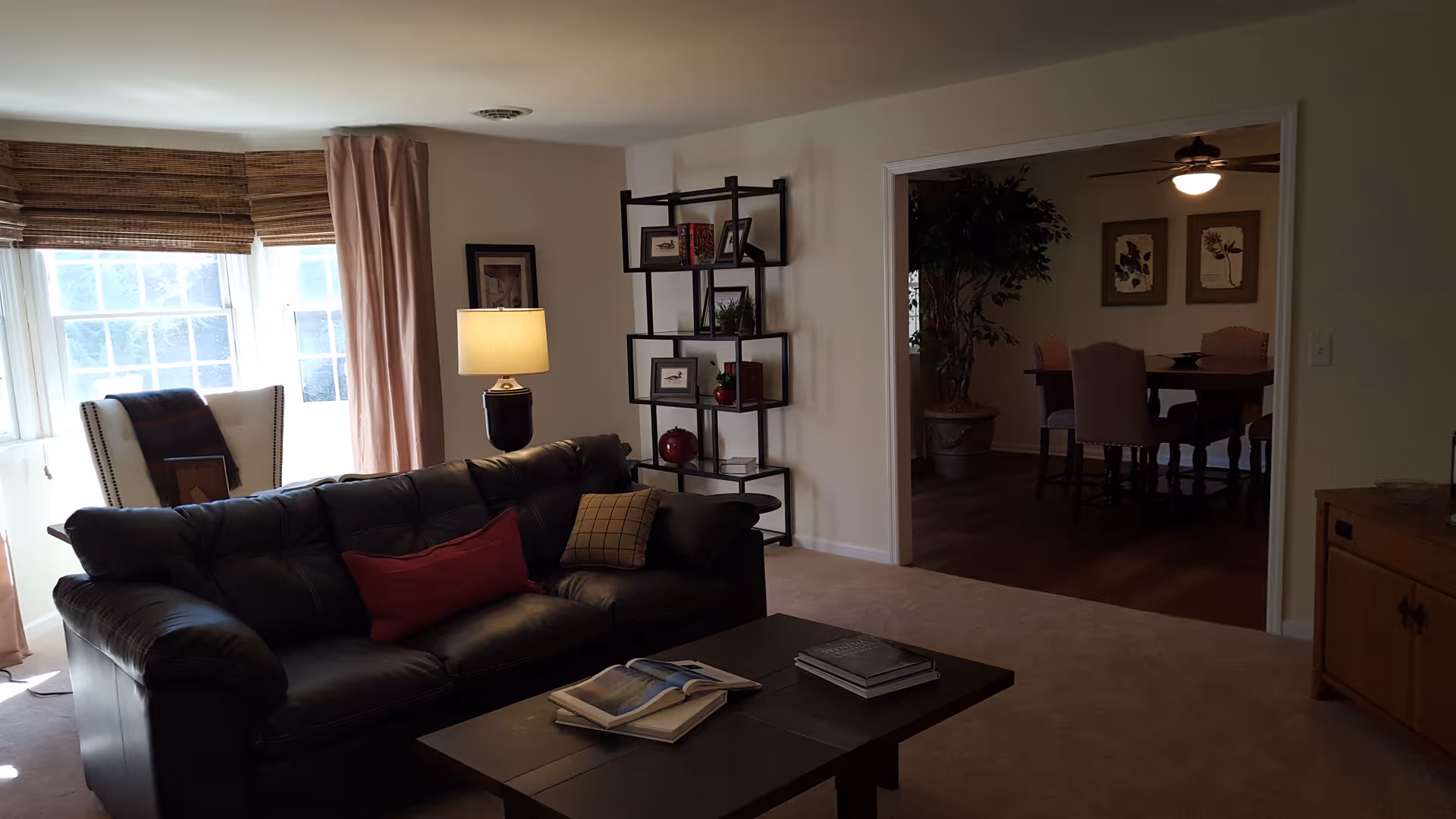 A cozy living room with a black leather sofa, coffee table, floor lamp, shelving unit, and a view into a dining room.