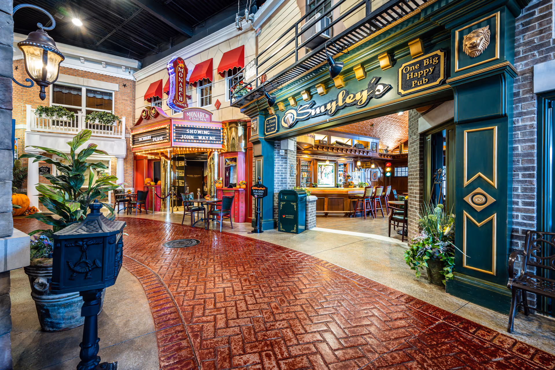 Indoor street scene in a senior living facility featuring a vintage-style Monarch Cinema with a marquee showing 'John Wayne' and a pub named Smyley's with a sign 'One Big Happy Pub'. The area has a brick-patterned floor, decorative plants, and warm lighting creating a cozy atmosphere.