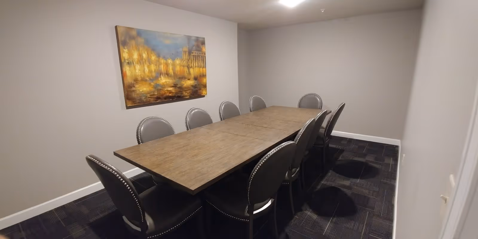 A small conference or meeting room with a rectangular wooden table surrounded by eight gray upholstered chairs with nailhead trim. The walls are painted light gray, and there is a large framed painting with warm yellow and blue tones hanging on one wall. The floor is covered with dark patterned carpet tiles.