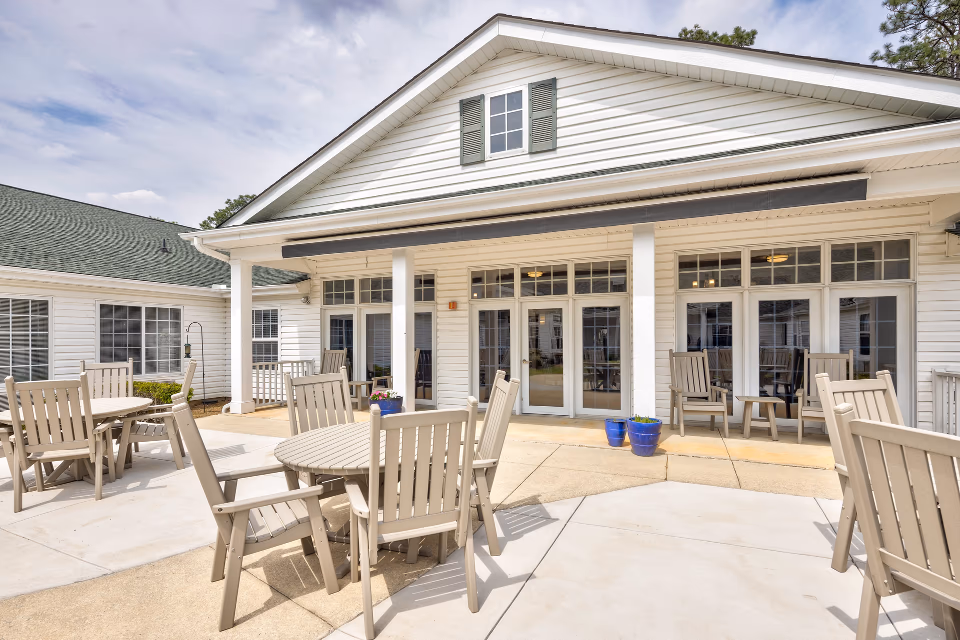 Outdoor patio area at TerraBella Southern Pines with multiple round tables and beige wooden chairs arranged on a concrete surface. The patio is adjacent to a white building with large windows and glass doors, and there are two blue flower pots near the entrance. The sky is partly cloudy.