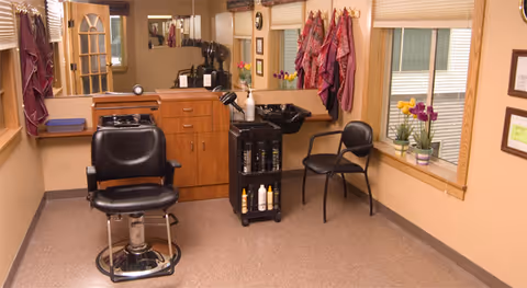 Small salon-style hairdressing room with two chairs, a shampoo sink, a cabinet, and windows with potted flowers.