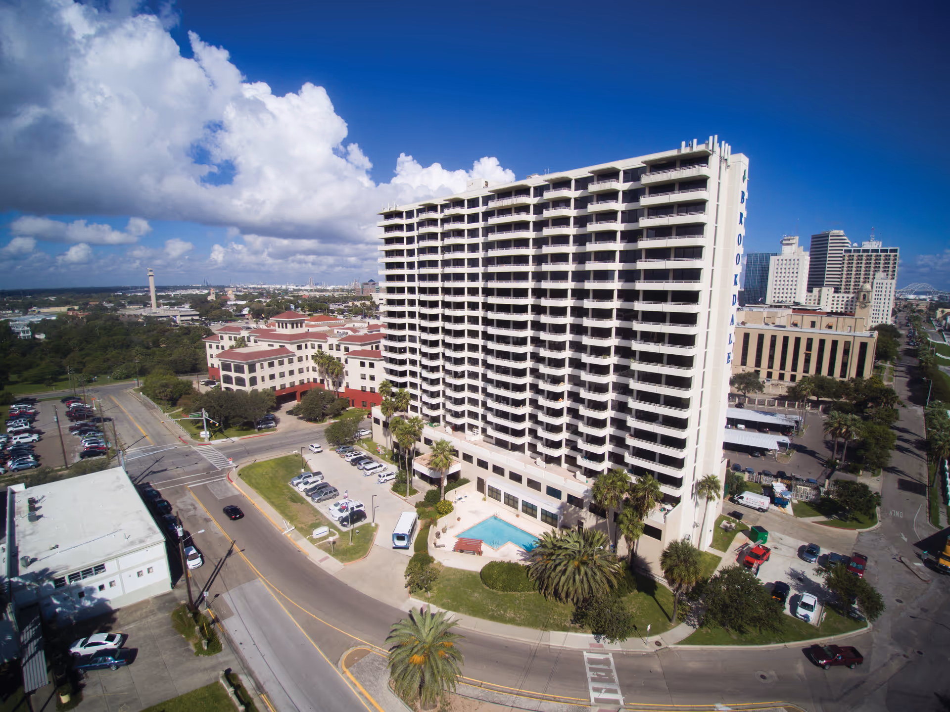 Aerial view of Brookdale Trinity Towers, a tall multi-story building with balconies, a pool and surrounding streets under a blue sky.