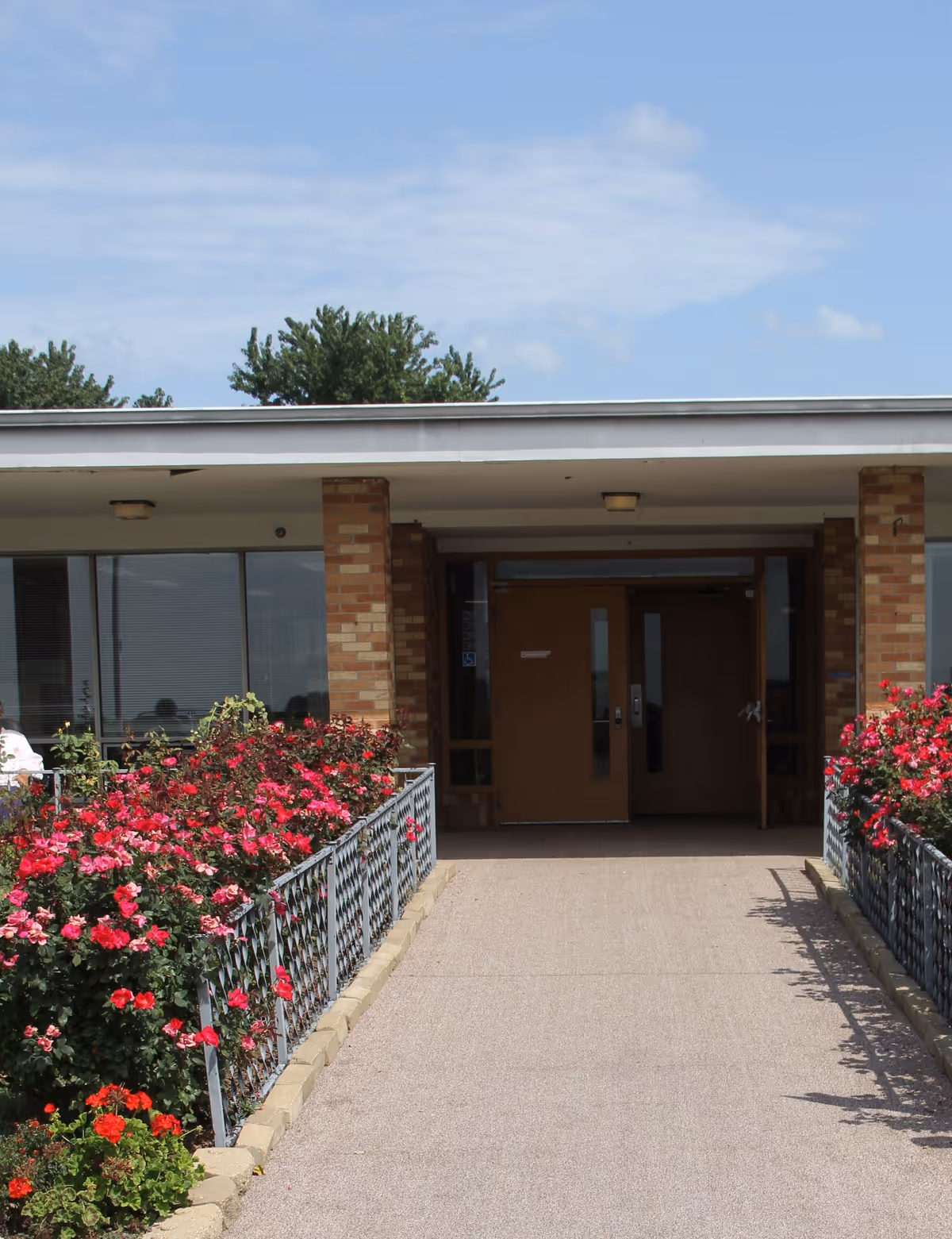 Entrance to a building with a pathway lined by metal railings and vibrant red and pink flowers on both sides. The building has brick pillars and large brown double doors under a flat roof. Trees and a blue sky with some clouds are visible in the background.